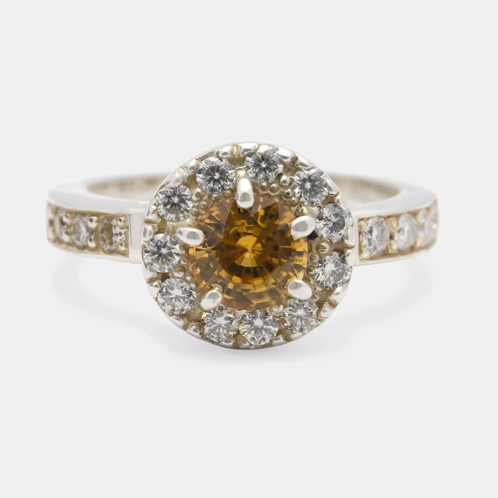 Anillo de plata visto de frente, con un circón anaranjado al centro rodeado por un halo de piedras blancas y más piedras engastadas en el aro.