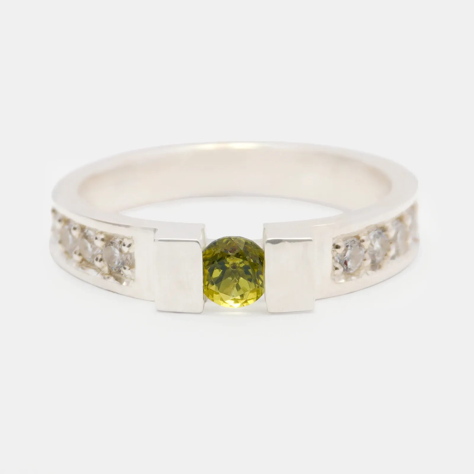 Anillo de plata visto de frente, con una piedra verde amarillenta sostenida entre los dos extremos del aro y piedras blancas engastadas a los costados.